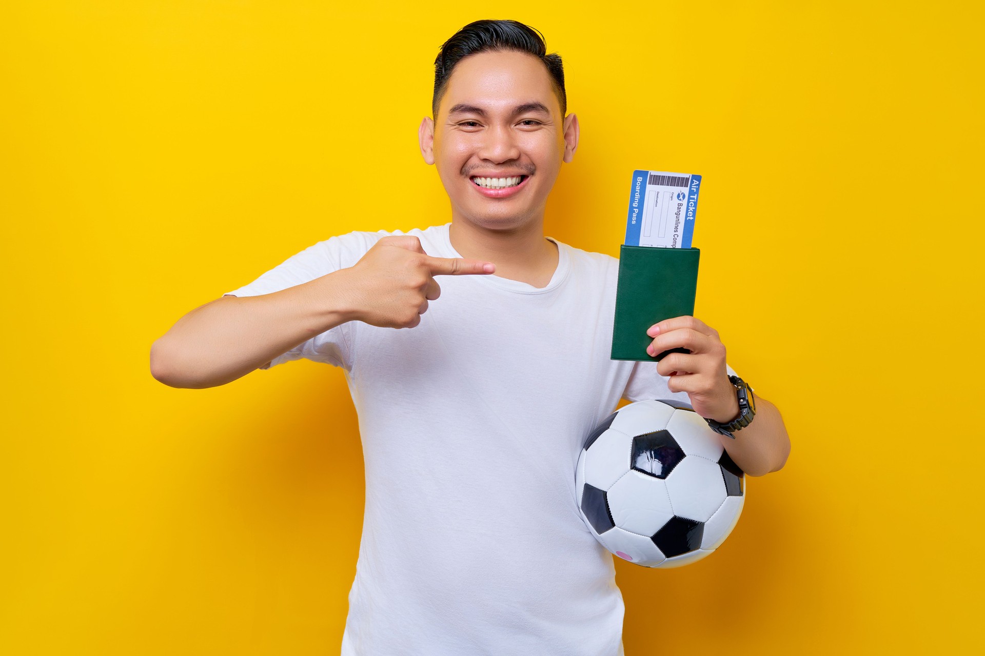 excited young Asian man football fan wearing a white t-shirt carrying a soccer ball and pointing a finger at a passport ticket boarding pass isolated on yellow background