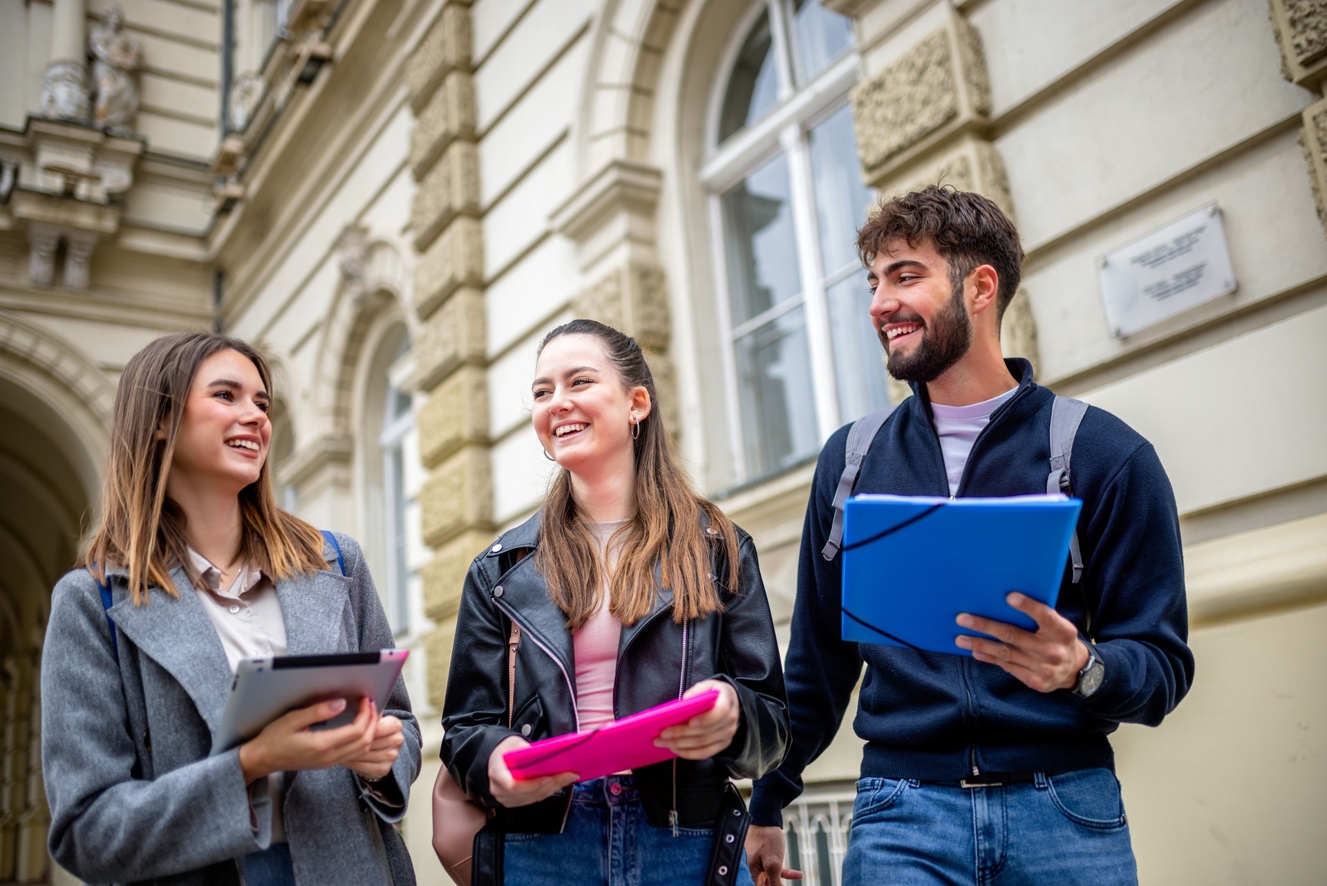 Three Students Holding Books Laughing and Talking Together Outdoors